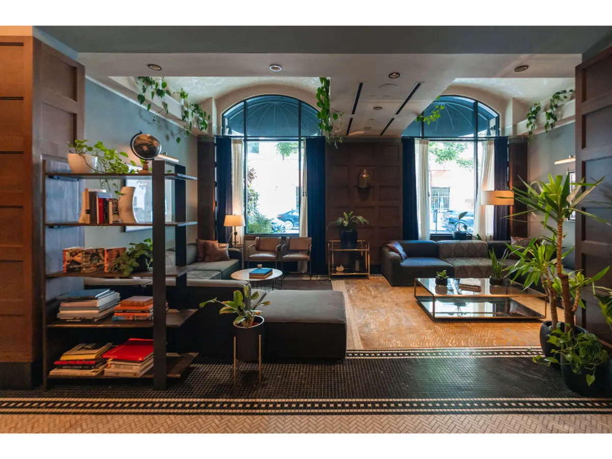 A sunlit lounge area with large windows, comfortable seating, a bookshelf with books and decorative items, and potted plants. The scene invites relaxation, reading, or casual work in a warm, welcoming atmosphere.