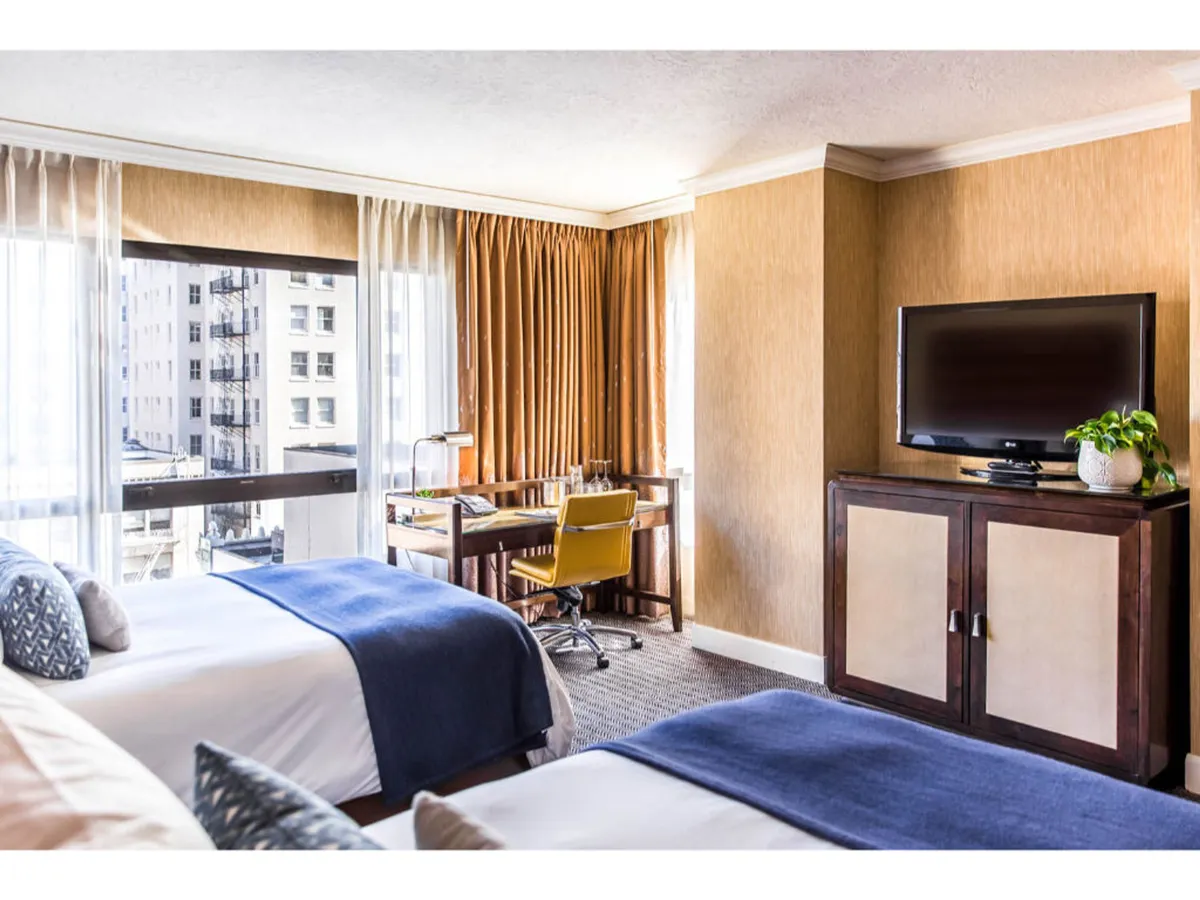 A contemporary main sleeping area with two double beds featuring blue throws, a stylish work desk with a yellow chair, a large window offering city views, and a flat-screen TV. The decor includes plush textiles and modern accents, capturing the essence of comfort and style.