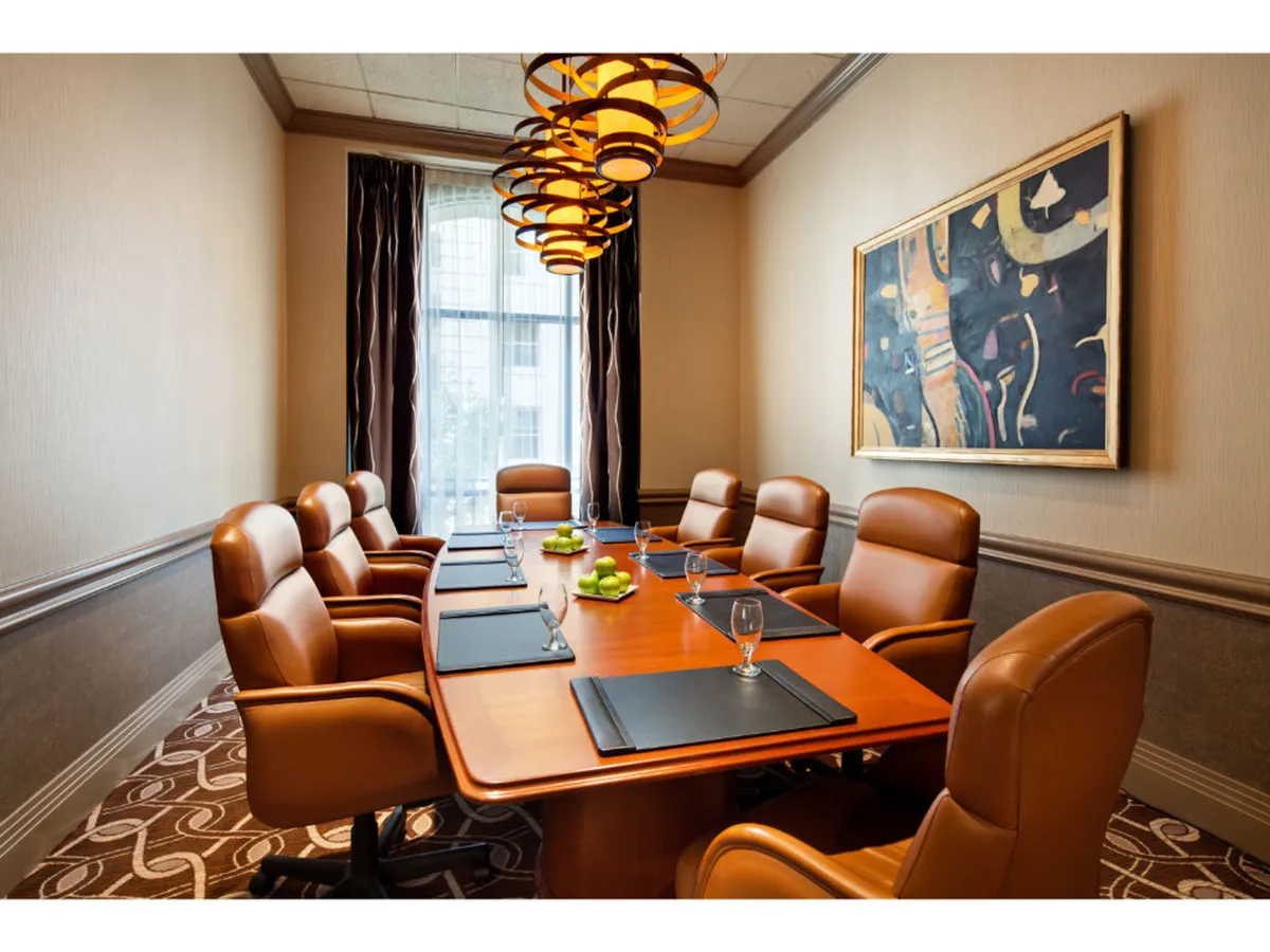 A spacious, sunlit conference room featuring a large wooden table surrounded by leather executive chairs. Decorative pendant lights hang above, and a piece of original artwork is prominently displayed on the wall. Light pours in through a window with sheer curtains, giving the room an inviting, creative ambiance.