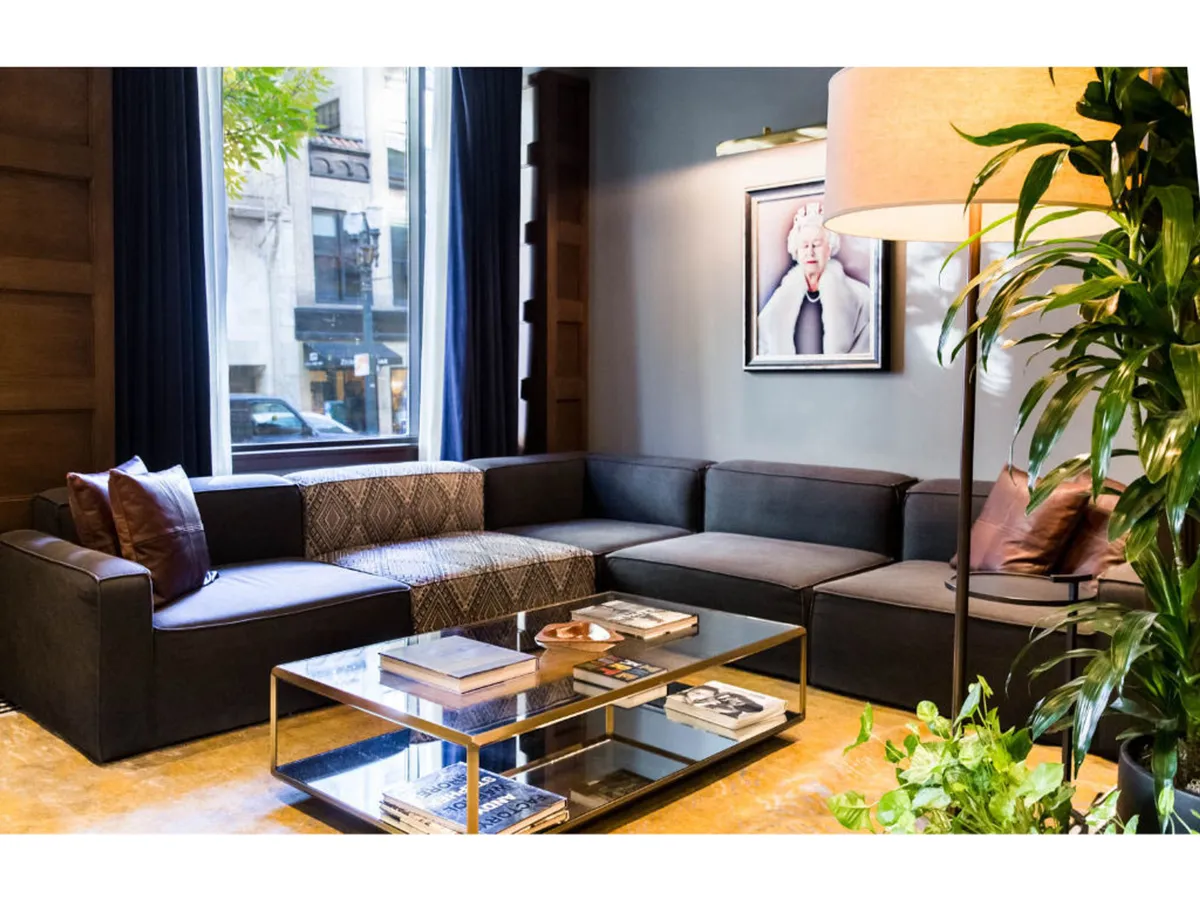 A contemporary communal lounge area with an L-shaped sectional sofa, glass coffee table, large windows offering city views, framed artwork, and a floor lamp. The space invites relaxation, casual conversation, and soaking in the local atmosphere.