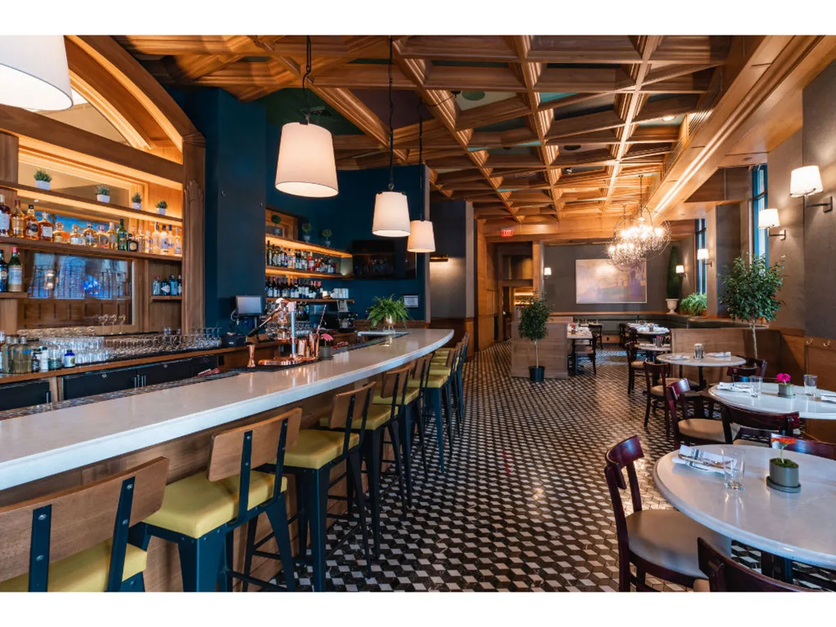 A stylish, warmly lit bar area with a sleek bar counter, hanging light fixtures, a wood-accented ceiling, and a classic checkered floor. The space is inviting and elegant, suggesting a perfect place to unwind after a workout or day of adventure.