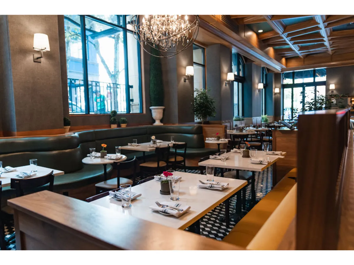 An elegant dining area with a striking chandelier, lush greenery, and a decorative wall. Tables and chairs are arranged for both intimate and group gatherings, creating a lively yet upscale environment. The scene captures guests dining, socializing, and celebrating, highlighting the communal and festive spirit.
