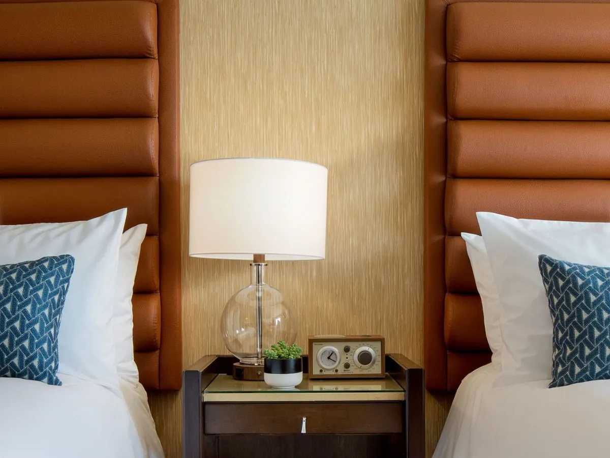 A cozy corner of the bedroom featuring a textured headboard, soft lighting from a bedside lamp, a nightstand with a decorative pillow, and a vintage-style radio. The ambiance is warm, inviting, and ideal for unwinding or starting the day with coffee.