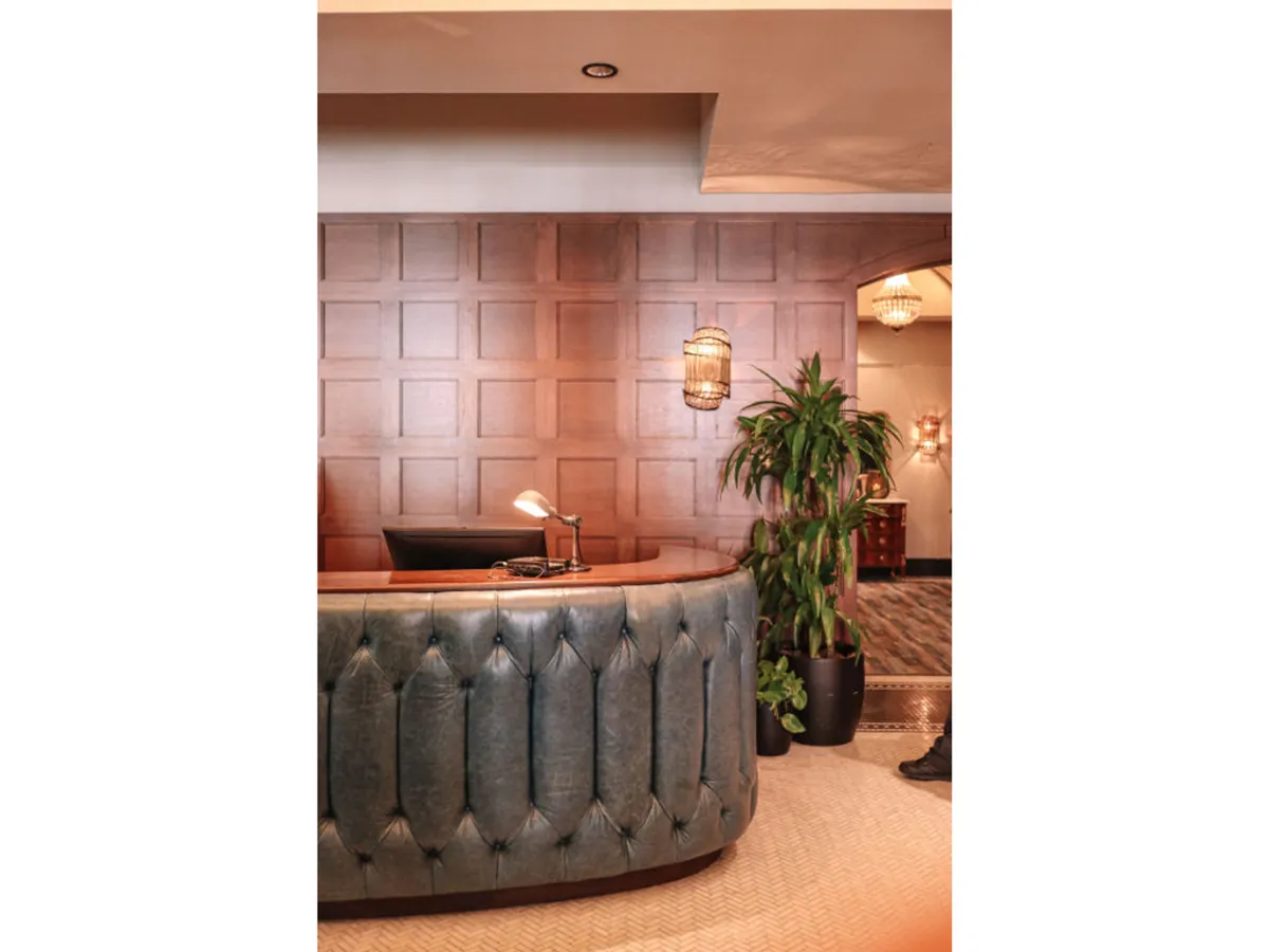 A sophisticated lobby scene featuring a curved reception desk, wooden paneled wall, hanging light fixtures, and a lush indoor plant. The environment feels modern and welcoming, ideal for arrivals and providing first impressions of service quality.