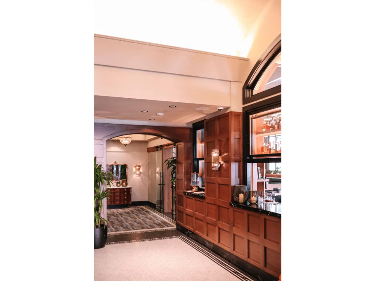 An upscale and inviting hotel bar area with wood paneling, a chandelier, decorative light fixtures, and lush plants. The space feels lively and welcoming, suitable for socializing or enjoying a welcome beverage upon arrival.