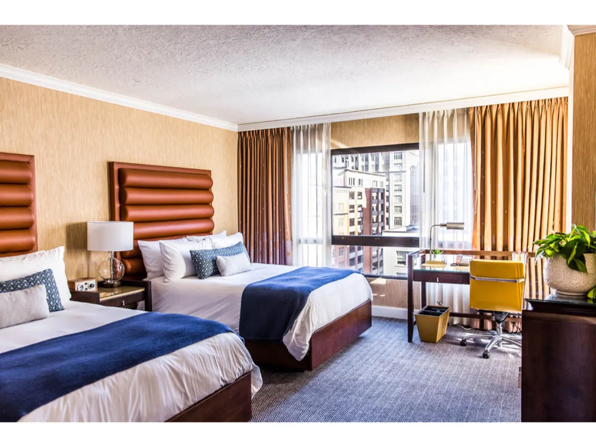 The image shows a stylish hotel room featuring two plush beds with unique decorative headboards, a modern desk area with a vibrant yellow chair, and a large window offering sweeping views of Portland’s cityscape. The room is bathed in natural light, with earthy tones and luxurious textures creating a welcoming, upscale ambiance.