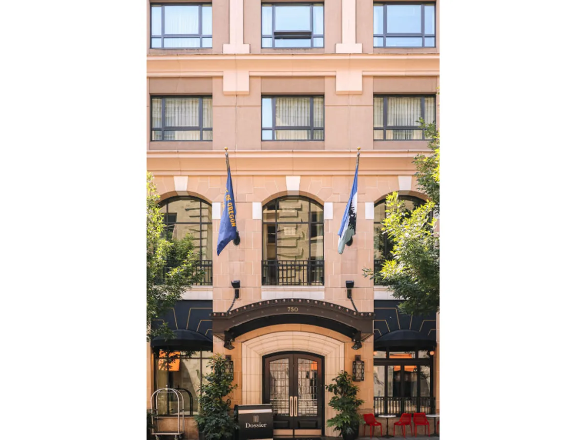 The photo spotlights the hotel’s elegant entrance with double doors, distinctive flags, outdoor seating, and architectural details. The mood is inviting and sophisticated, with glimpses of people enjoying a coffee and the energetic street scene nearby.