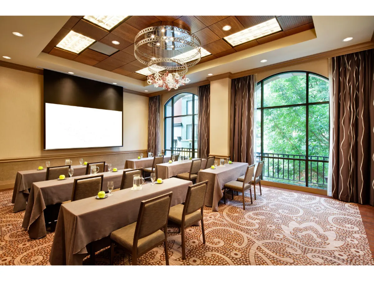 The image features a well-lit business meeting room with large windows letting in natural light, an elegant chandelier, a presentation screen, and a decorative carpet. The environment appears both focused and relaxed, suitable for workshops, presentations, or informal networking.